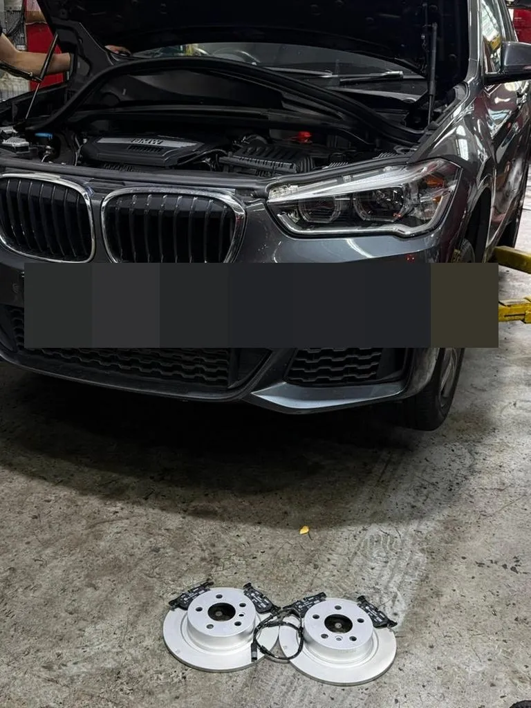 New rear brake discs, pads and wear sensor laid out in front of a BMW X1 at our Singapore workshop