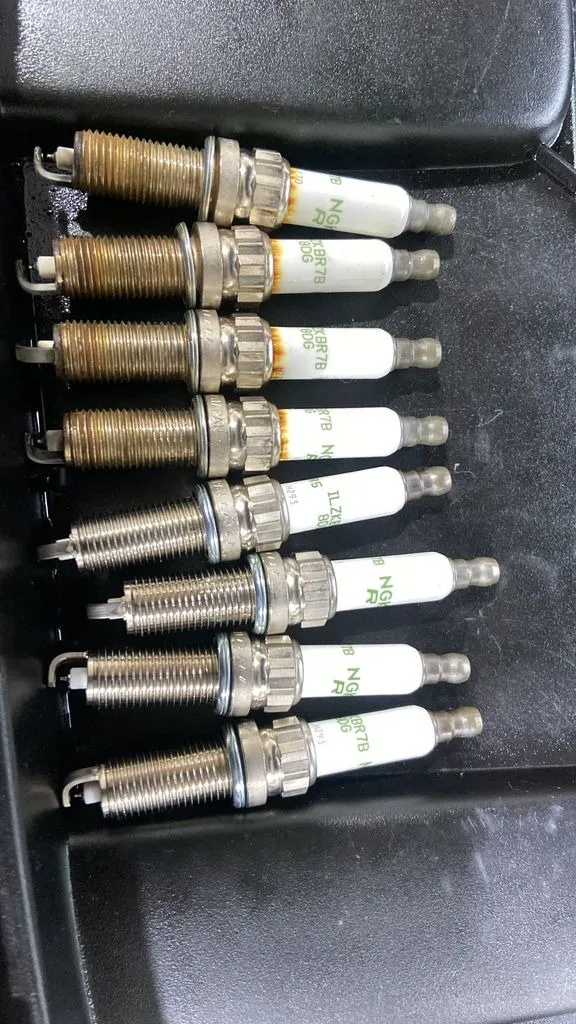 Four old spark plugs next to four new NGK spark plugs from a Peugeot 5008 service